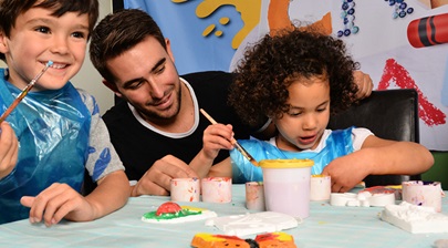 Two kids painting pottery whilst being supervised by a man