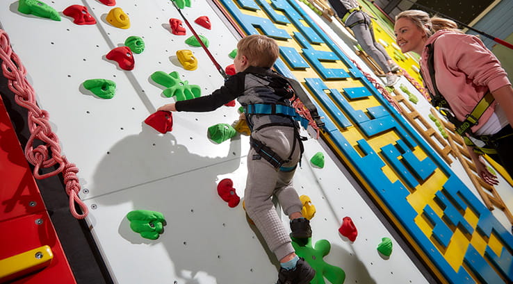 A young boy scaling the indoor climbing wall whilst his mother watches