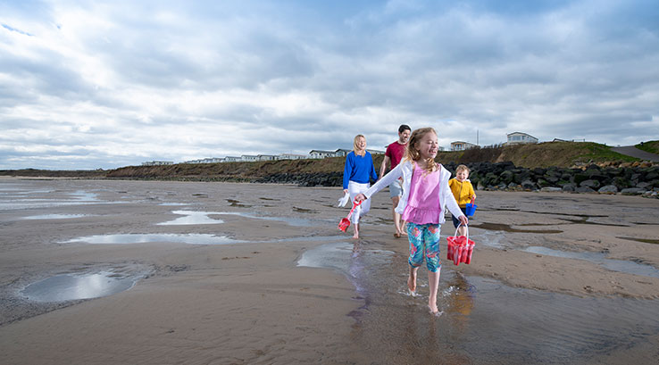 Two kids walking along a sandy beach with buckets and spades whilst parents walk behind