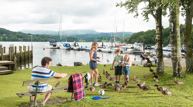 A family feeding ducks on the grass by the edge of Lake Windermere